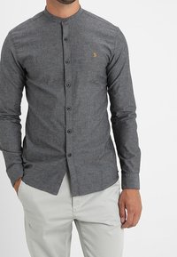 Chemise boutonnée en tissu gris foncé, avec un design sans col, des manches longues et un petit logo doré sur la poitrine. Texture lisse.