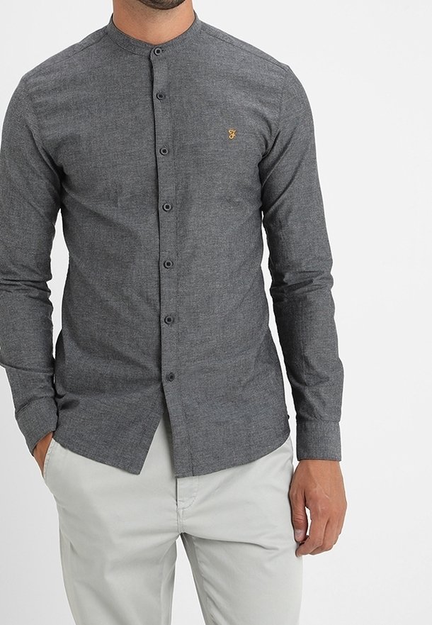 Chemise boutonnée en tissu gris foncé, avec un design sans col, des manches longues et un petit logo doré sur la poitrine. Texture lisse.