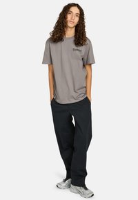 Persona joven con cabello largo y rizado, vistiendo una camiseta gris de Element, pantalones negros y zapatillas plateadas, de pie con las manos en los bolsillos.