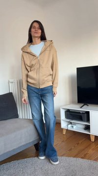 Beige hettejakke med glidelås over en lyseblå skjorte, kombinert med vide blå jeans og blå joggesko. Glatt stoff og avslappet design.