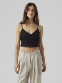 Crni mrežasti crop top s podesivim trakama i valovitim rubom, uparen s svijetlo sivim hlačama visog struka.