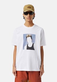 Biała bawełniana koszulka z graficznym nadrukiem twarzy na niebieskim tle. Noszona z beżową czapką i czarnymi okularami przeciwsłonecznymi.