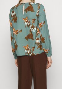 Blouse à manches longues en tissu teal présentant des motifs floraux et d'oiseaux en orange et blanc, avec un détail d'ouverture en forme de clé au dos et des poignets boutonnés.