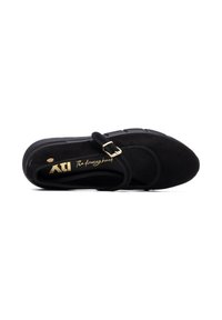 Zapato negro de ante tipo slip-on con puntera redondeada, correa ajustable con hebilla dorada y suela texturizada. Marca visible en letras doradas.