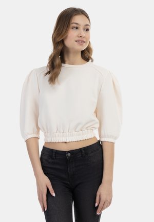 Felpa corta di un beige chiaro con maniche a sbuffo e vita elastica, abbinata a jeans skinny scuri. Tessuto dalla consistenza liscia.