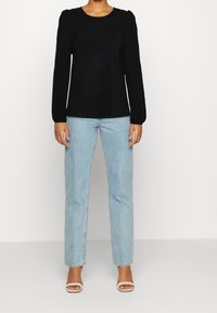 Maglia nera a maniche lunghe con spalle arricciate, abbinata a jeans dritti azzurri e sandali bianchi con cinturino. Tessuto uniforme in tutto.