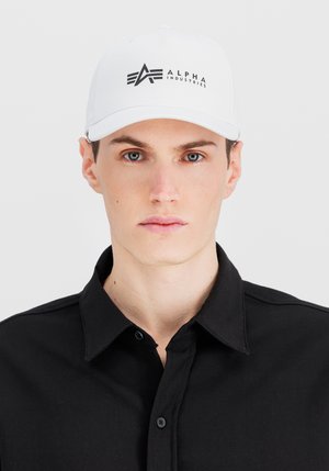 Valkoinen lippalakki, jossa on jämäkkä muotoilu; siinä on musta logo, jossa lukee "ALPHA INDUSTRIES" ja tyylikäs, kaareva lippa. Musta paita alla.