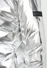 Casaco puffa prateado com acabamento brilhante, apresentando textura franzida, detalhe com fecho éclair e uma etiqueta de borracha preta.