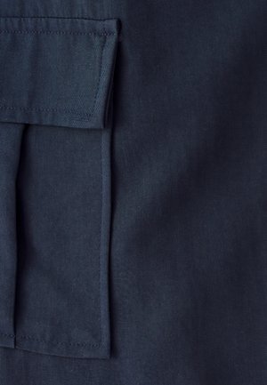 Mere lähedalt vaadatav tumesinine kangas, millel on ristkülikukujuline klapiga tasku korralike õmblustega servades.