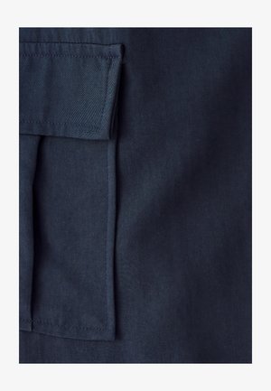 Lähikuva laivastonsinisestä kankaasta, jossa on suorakaiteen muotoinen läppätasku siisteillä saumoilla reunoilla.