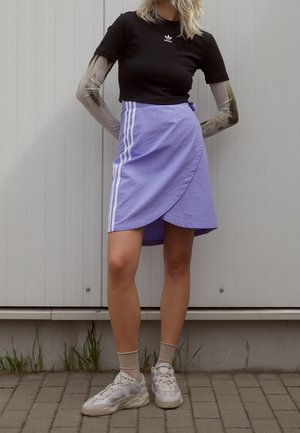 Zwarte crop top met wit logo, lavendelkleurige wikkelrok met witte zijstrepen en beige sneakers. De achtergrond heeft een lichtgrijze muur.