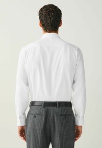 Biela košeľa s dlhým rukávom a hladkou textúrou, nosená s tmavými nohavicami s tenkými pásmi. Pohľad zozadu ukazuje lem košele a priliehavý vzhľad.