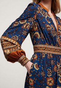 Blå blommig klänning med långa, utsvängda ärmar och mönstrade manschetter. Har en insydd midja och sidofickor. Rik på textur och färg.