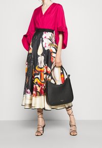 Donna che indossa un blusa rosa brillante e una gonna midi a stampa astratta colorata, mentre tiene in mano una borsa nera, in piedi su sandali con tacco neri e fasciati.