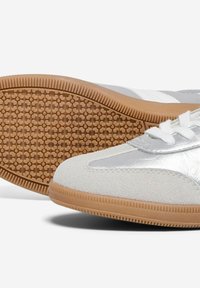 Baskets de sport blanches et argentées avec une semelle en caoutchouc texturé et des détails en daim, dotées d'une épaisse semelle en gomme et d'un système de laçage classique.