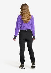 Kvinna med långt, vågigt hår som bär en lila fleece-tröja, svarta utem trousers och vandringsskor, stående med ryggen mot kameran mot en vit bakgrund.