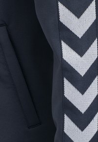 Navy urheiluhousuissa on teksturoitu kangas, jossa on valkoinen sauma-kuvio sivuilla. Vasemmassa lahkeessa on piilotasku.