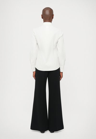 Biela košeľa s dlhým rukávom a jednoduchým golierom, skombinovaná s čiernymi voľnými nohavicami s vysokým pásom, ktoré majú hladkú textúru a strih prispôsobený postave.