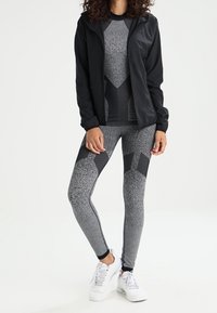 Giacca nera con zip sopra una maglietta grigia a maniche lunghe con motivo e leggings abbinati. L'outfit presenta dettagli in rete e una vestibilità slim. Sneakers bianche completano il look.