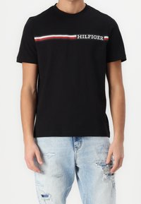Osoba oblečená v černém tričku s krátkým rukávem, které má vodorovný pruh v červené, bílé a modré barvě a nápis "HILFIGER" na prsou.