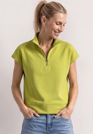 Kvinde med blond hestehale iført limegrøn kortærmet trøje med lynlås og blå jeans, smilende mens hun kigger nedad med hænderne i lommerne.