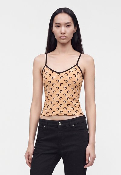 Jeune femme aux longs cheveux noirs portant un caraco pêche avec des motifs en forme de croissant de lune noirs et un pantalon noir sur un fond uni.