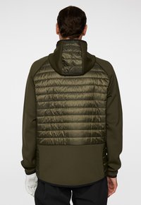 Man som bär en mörkgrön vadderad jacka med huva, svarta byxor och vita handskar, vänd bort från en enfärgad bakgrund.