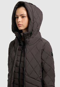 Young woman wearing a dark gray quilted hooded jacket with two layered front zippers, facing left on a gray background.
