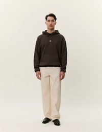 Felpa marrone con tasca frontale e cordino, abbinata a pantaloni avorio larghi e scarpe nere. Dettaglio logo semplice sulla felpa.
