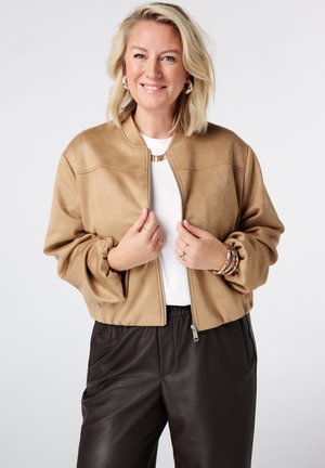 Glimlachende vrouw met blond haar die een beige jas draagt over een witte top en zwarte broek, met beide handen de jasrits vasthoudend.