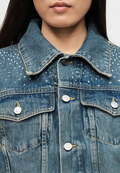 Νεαρό άτομο που φοράει μπλε τζιν μπουφάν με ασημένια καρφιά στους ώμους και κουμπωμένο μπροστά, με εμφανές το γιακά και το πάνω μέρος του στήθους.