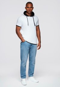 Muž stojící v bílém krátkém rukávu s černým lemem, světlých modrých džínách a bílých teniskách na jednobarevném pozadí.