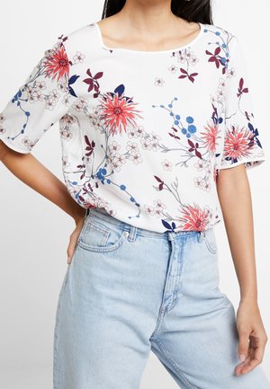 Kortærmet hvid bluse med levende blomsterprint i rød, blå og lilla. Har en rund halsudskæring og en løs pasform. Sat sammen med lyseblå jeans.