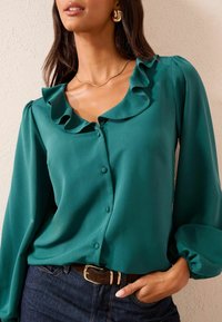 Teal blouse with ruffled neckline, long sleeves, and button front. Made of smooth fabric, features elastic cuffs and a subtle sheen.