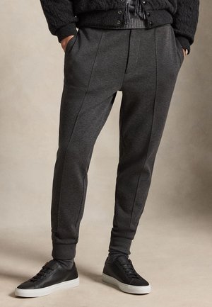 Persoon die donkergrijze taps toelopende joggerbroeken met zijzakken draagt, zwarte sneakers met witte zolen en een zwarte gestructureerde trui.