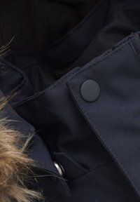 Mörkblå tygjacka inuti med pälsdetalj, med tryckknapp och kontrasterande sömmar. Mjuk textur synlig i tygvikarna.