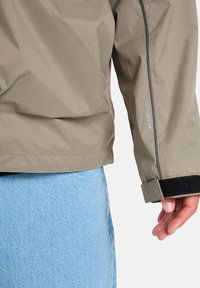 Lichtgewicht, khaki regenjack met een gestructureerde afwerking, elastische manchetten en een "DRYFLIGHT" logo. Beschikt over een strak ontwerp zonder zichtbare patronen.