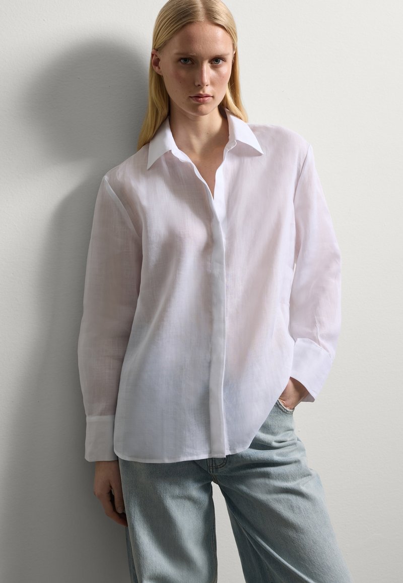 Camicia bianca in lino con bottoni, maniche lunghe e colletto classico, abbinata a jeans in denim blu chiaro. Tessuto morbido e vestibilità rilassata.