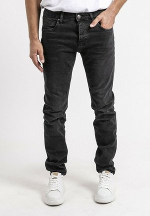 Homme portant un jean noir slim et des baskets blanches, une main dans la poche, sur fond blanc.