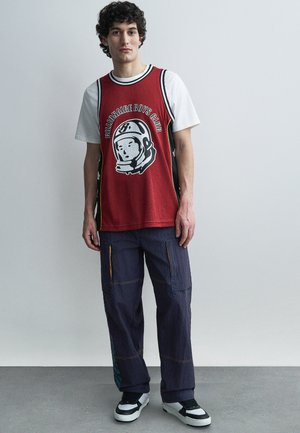 Jaunas vyras su garbanotais plaukais, dėvintis raudoną krepšinio marškinėlį ant baltos marškinėlių, laisvus tamsius krovinius kelnes ir juodai baltus sportbačius, stovintis patalpose.