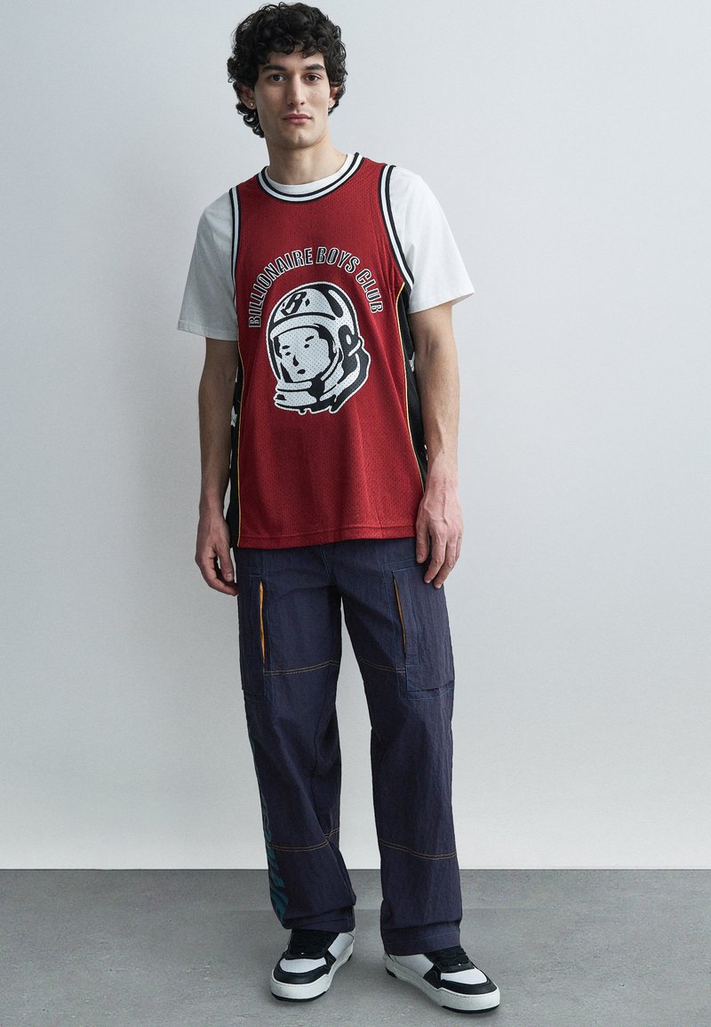 Νέος με σγουρά μαλλιά που φοράει μια κόκκινη φανέλα μπάσκετ πάνω από λευκή μπλούζα, άνετο σκούρο παντελόνι cargo και ασπρόμαυρα αθλητικά παπούτσια, στέκεται μέσα σε εσωτερικό χώρο.