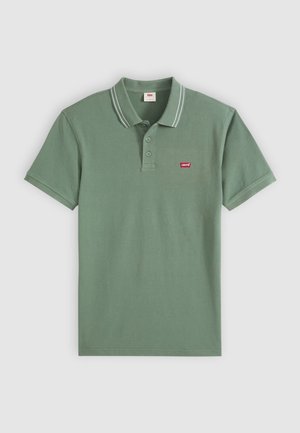 Grønn bomullspolo med klassisk krage, to knapper, korte ermer og hvite aksentstriper. Har et lite rødt logo-patch.