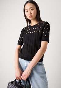 Black ribbed knit top featuring circular cutouts on the shoulders and sleeves, paired with high-waisted light blue denim jeans.