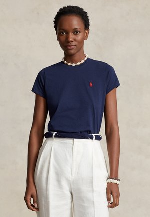 Vrouw die een marineblauw T-shirt met een rood logo draagt, witte hoge taillebroek, schelpenketting en gevlochten armbanden, staande tegen een effen achtergrond.
