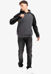 Felpa grigia con maniche nere, logo testurizzato sul davanti, tasca a marsupio, abbinata a jeans scuri e sneakers bianche.