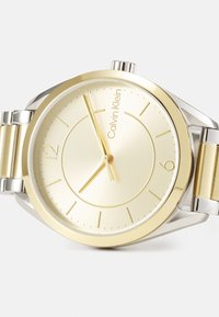 Reloj de pulsera de metal dorado y plateado con un diseño minimalista, que cuenta con una esfera de un dorado claro, marcadores de horas delgados y una correa suave.