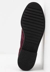 Semelle en caoutchouc noire avec des rainures horizontales et un talon texturé sur une chaussure en daim rouge foncé, inclinée contre une surface blanche.