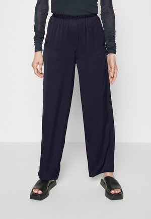 Marinblå vida byxor med elastisk midja, tillverkade av ett mjukt tyg. Matchas med kraftiga svarta sandaler med fyrkantig tå och platt sula.