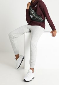 Felpa bordeaux con logo, pantaloni della tuta grigi e scarpe da ginnastica bianche. Borsa a tracolla in camo nero e grigio indossata sul torso. Texture lisce, vestibilità comoda.