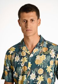 Camisa de manga corta con un patrón tropical de limones amarillos y flores blancas sobre un fondo verde oscuro. Botonadura en la parte delantera con cuello.